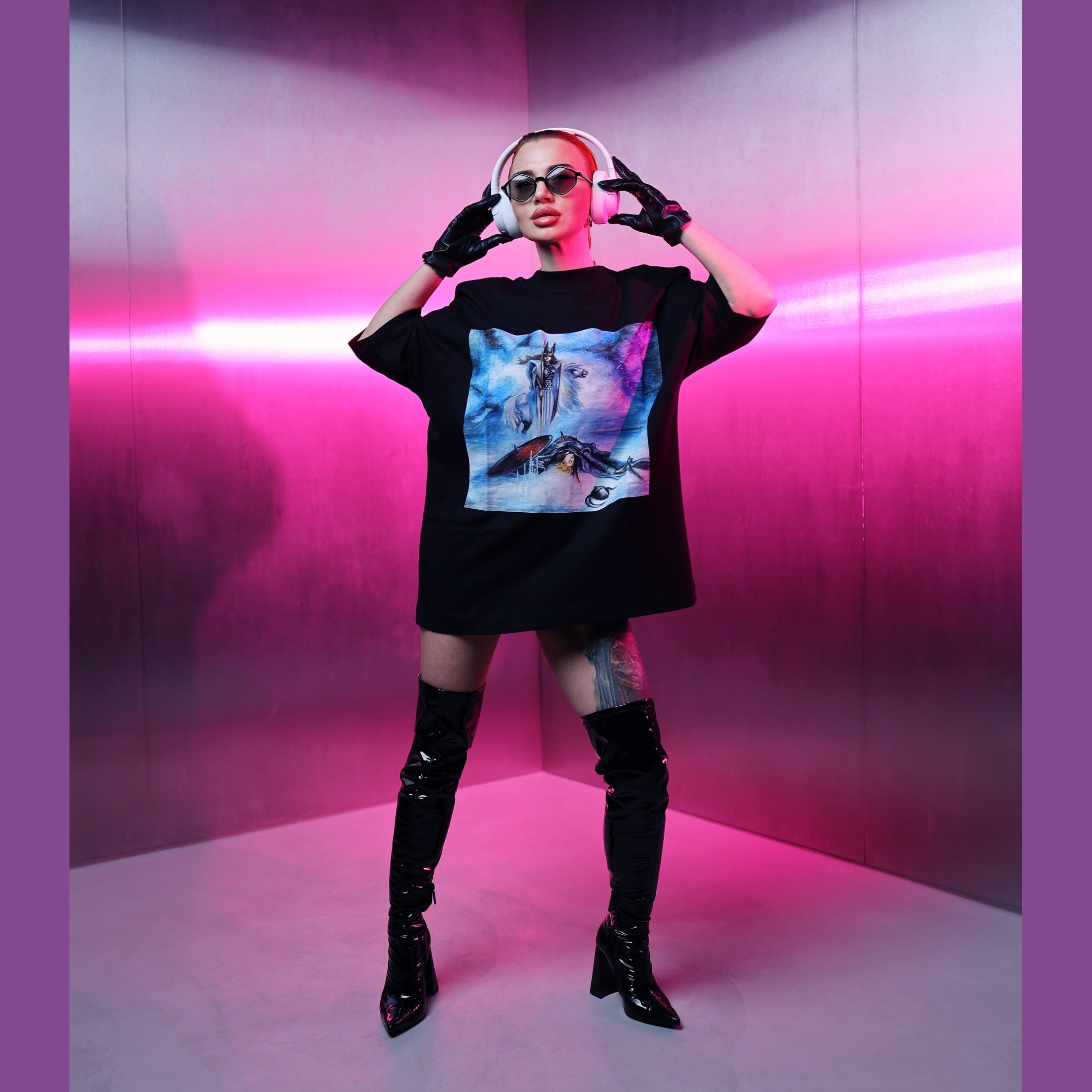 Person wearing a black oversized t-shirt with a colorful graphic design, black knee-high boots, and sunglasses in a pink-lit room.