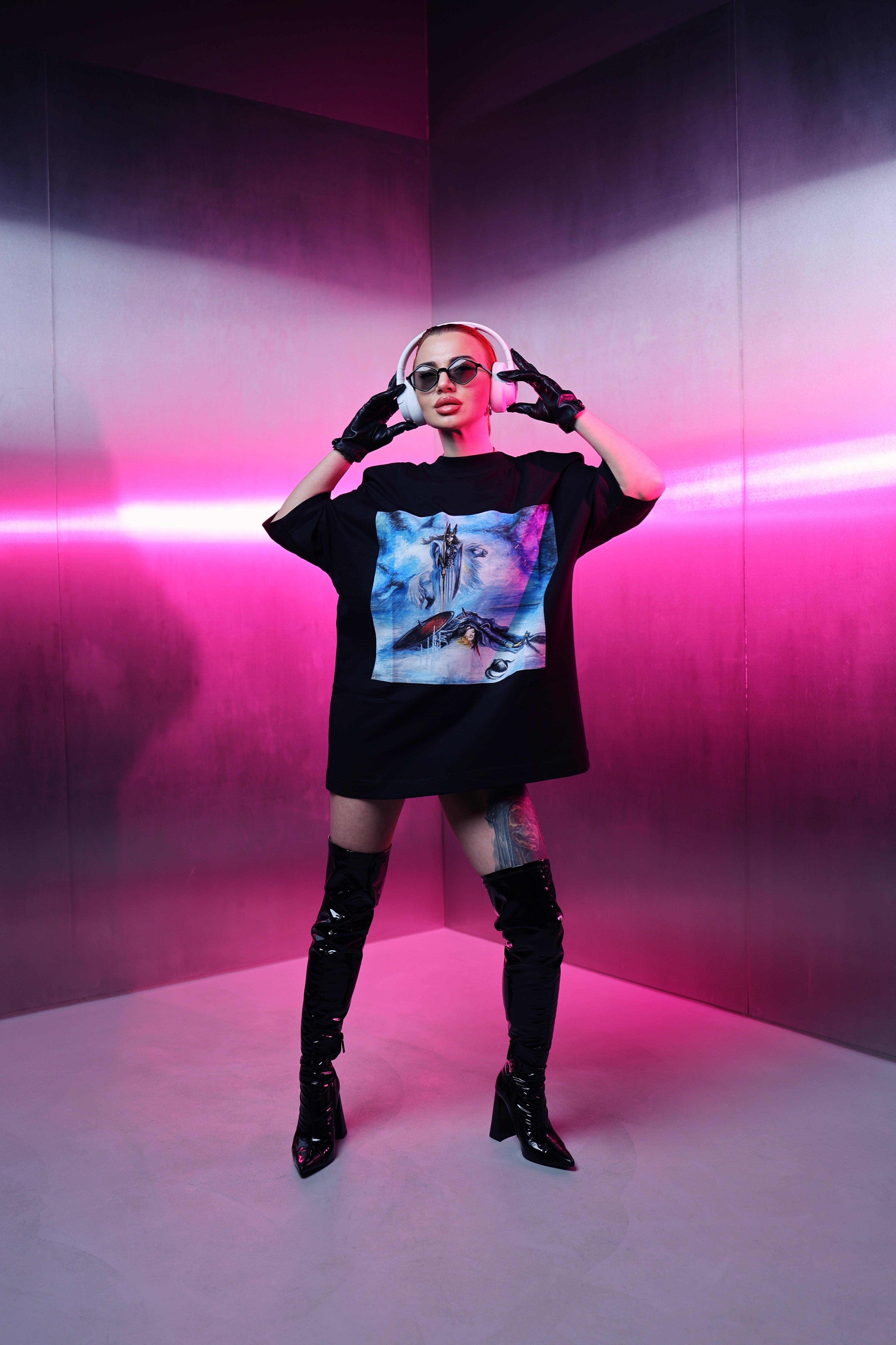 Person wearing a black oversized t-shirt with a colorful graphic design, black knee-high boots, and sunglasses in a pink-lit room.