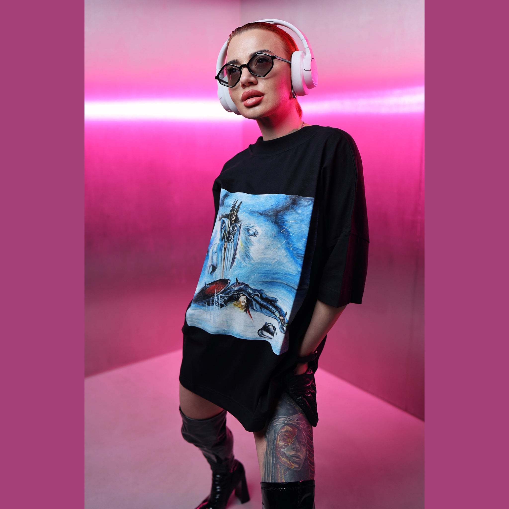 Person wearing a black oversized shirt with a graphic design, white headphones, and black sunglasses in a pink-lit room.